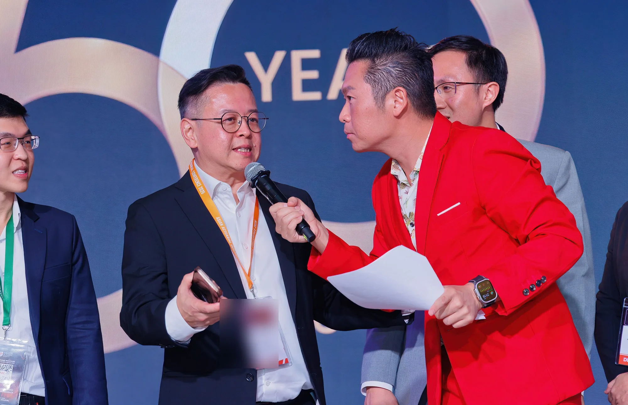 James Yang on stage with a microphone in a red suit, hosting the SAS 50th Anniversary Gala at the Asian Actuarial Conference 2026