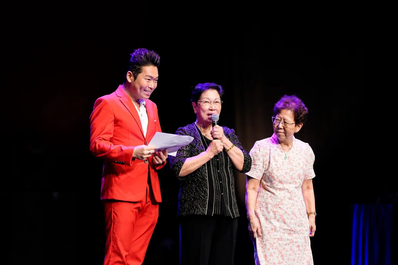 James Yang in a red suit on stage with two guests at the microphone